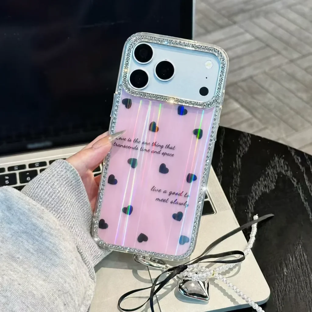 Pink iridescent heart phone case with a crystal-style border and matching camera surround, held in front of a laptop